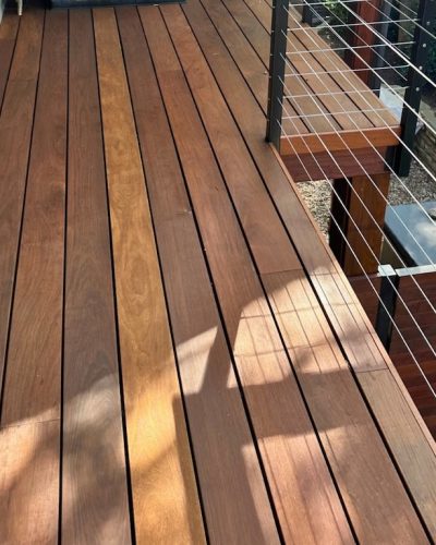 Wooden deck with warm-toned planks, sunlight casting soft shadows. Modern metal railing on the right with horizontal cables, garden visible below.