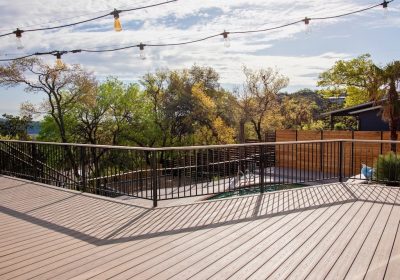 Expansive wooden deck with string lights overhead, black railing casting shadows, surrounded by leafy trees and a fenced area, under a partly cloudy sky.