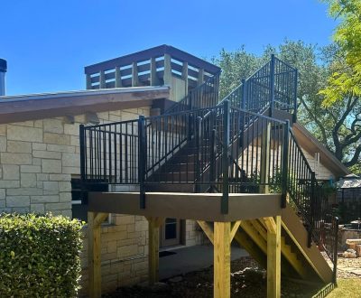 Outdoor staircase with black metal railings leading to a rooftop deck on a stone house. The setting is sunny with trees and an umbrella in the background.