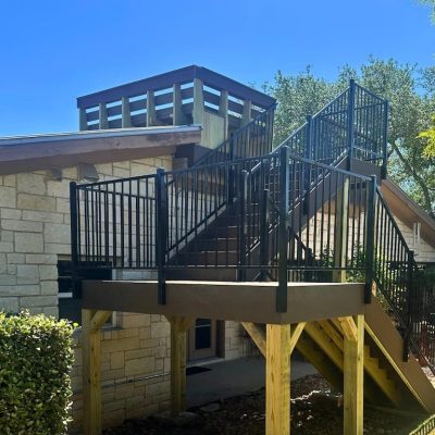 Outdoor staircase with black metal railings leading to a rooftop deck on a stone house. The setting is sunny with trees and an umbrella in the background.