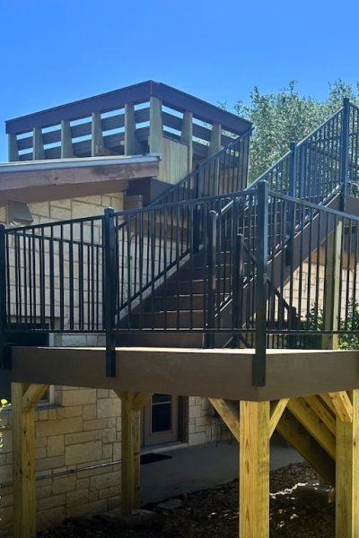 Outdoor staircase with black metal railings leading to a rooftop deck on a stone house. The setting is sunny with trees and an umbrella in the background.