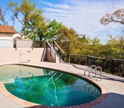 Outdoor pool area with oval-shaped pool surrounded by a concrete deck, brick trim, and metal railings. Trees and a house in the background under a sunny sky.