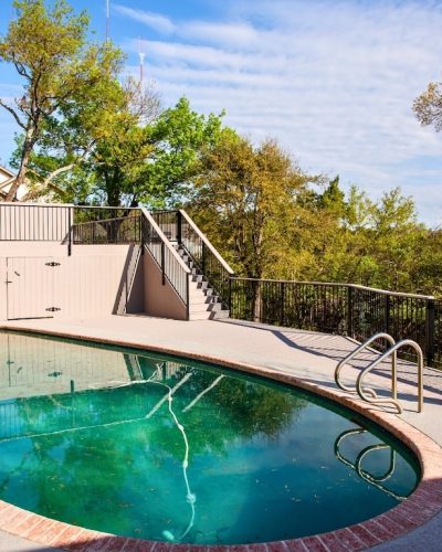 Outdoor pool area with oval-shaped pool surrounded by a concrete deck, brick trim, and metal railings. Trees and a house in the background under a sunny sky.