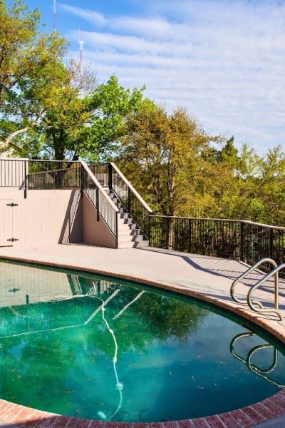 Outdoor pool area with oval-shaped pool surrounded by a concrete deck, brick trim, and metal railings. Trees and a house in the background under a sunny sky.