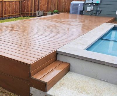 Wooden deck with steps beside a modern pool, surrounded by a wooden fence, creating a serene outdoor space. The finish is glossy and wet from rain.