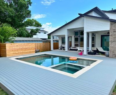 Modern backyard with a rectangular pool, surrounded by a gray deck. The pool has colorful floating toys. A large, white house with a patio is in the background. Vibrant green trees and a wooden fence frame the scene, conveying a peaceful, leisurely atmosphere.