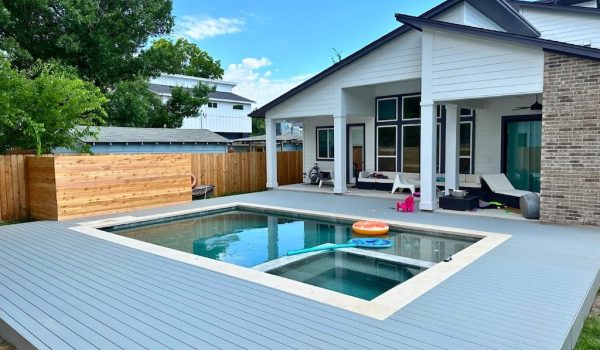 Modern backyard with a rectangular pool, surrounded by a gray deck. The pool has colorful floating toys. A large, white house with a patio is in the background. Vibrant green trees and a wooden fence frame the scene, conveying a peaceful, leisurely atmosphere.