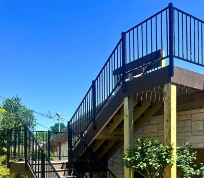 Outdoor wooden staircase with black railings against a stone building, surrounded by lush green trees under a clear blue sky.