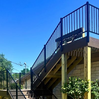Outdoor wooden staircase with black railings against a stone building, surrounded by lush green trees under a clear blue sky.