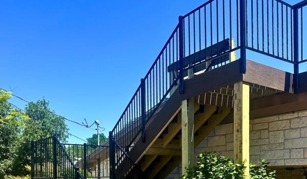 Outdoor wooden staircase with black railings against a stone building, surrounded by lush green trees under a clear blue sky.