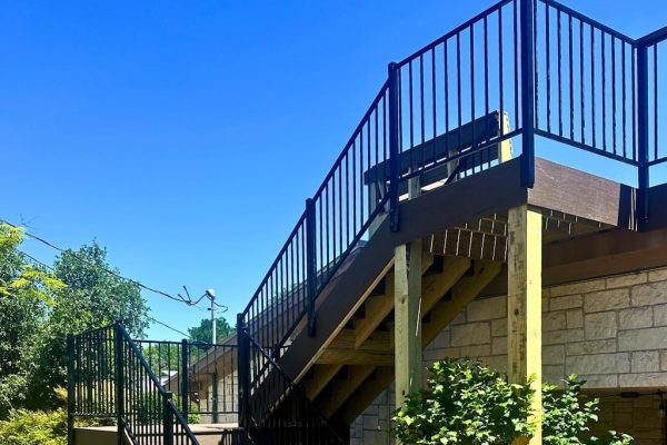 Outdoor wooden staircase with black railings against a stone building, surrounded by lush green trees under a clear blue sky.