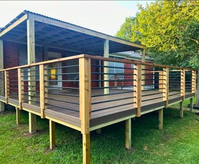 Wooden deck with metal and wood railing attached to a brick house. The deck is elevated with a corrugated metal roof and set against a grassy yard and trees.