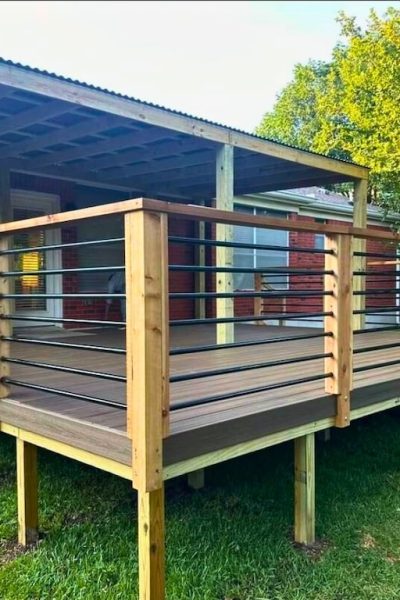 Wooden deck with metal and wood railing attached to a brick house. The deck is elevated with a corrugated metal roof and set against a grassy yard and trees.