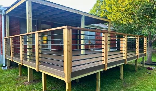 Wooden deck with metal and wood railing attached to a brick house. The deck is elevated with a corrugated metal roof and set against a grassy yard and trees.