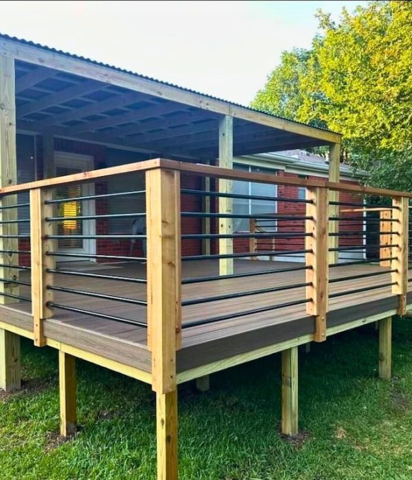 Wooden deck with metal and wood railing attached to a brick house. The deck is elevated with a corrugated metal roof and set against a grassy yard and trees.